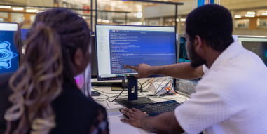 Two computer science students sitting at a computer. One student is showing the other something on the monitor by pointing at it