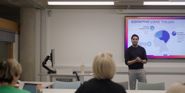 A lecturer teaching at the front of a classroom while students watch and listen