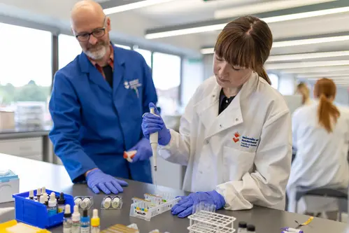 A student dressed in a white lab coat and protective eyewear and gloves using a pipette in a laboratory, while a member of staff looks on