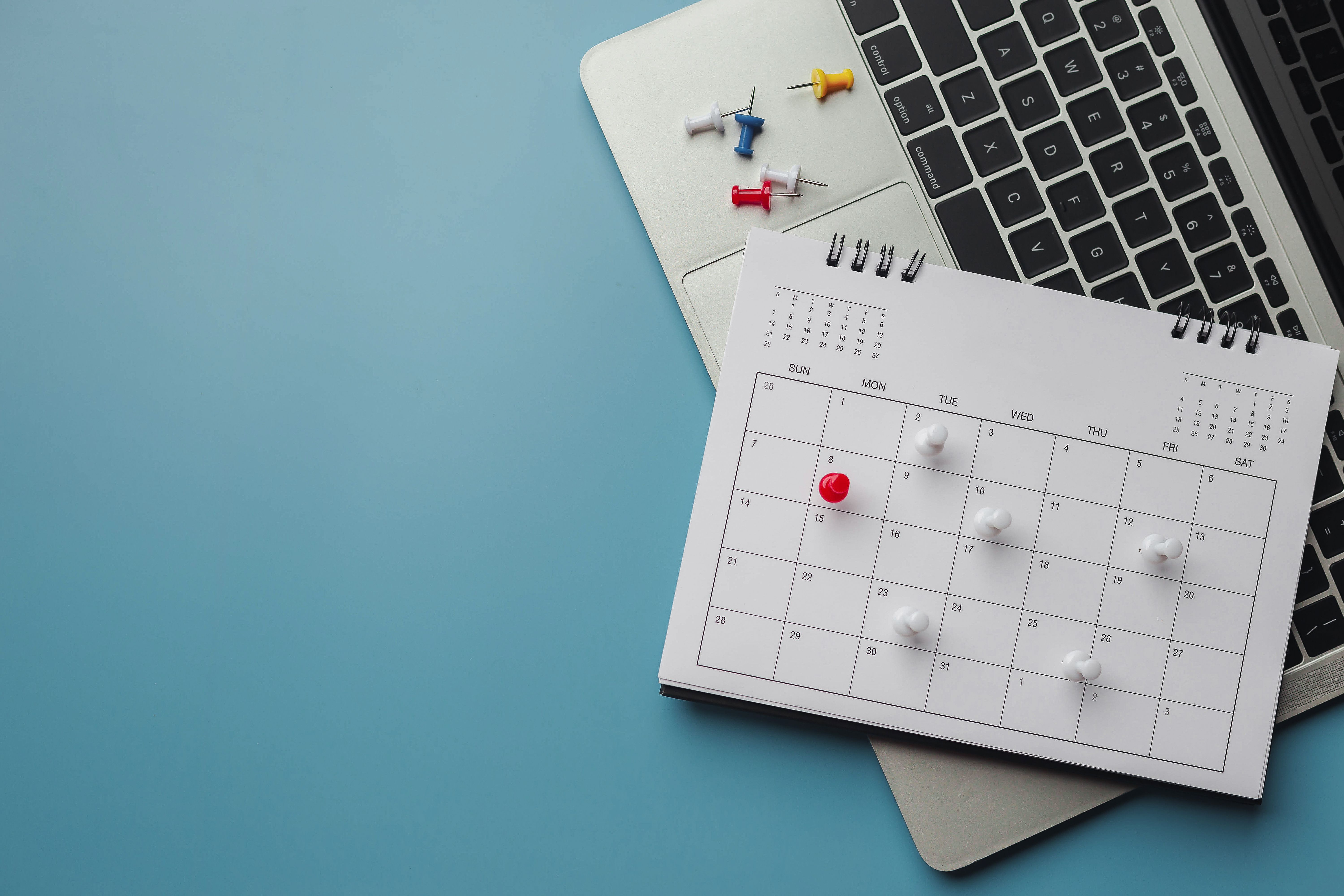 A laptop to the right of the image with monthly calendar on top. The calendar has coloured pins in it.