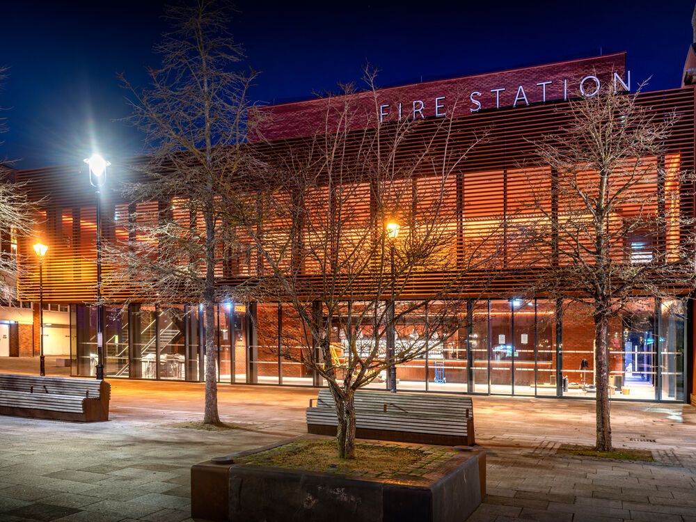 A view of the Fire Station bar and restaurant at night. The building is lit up with trees and benches outside.