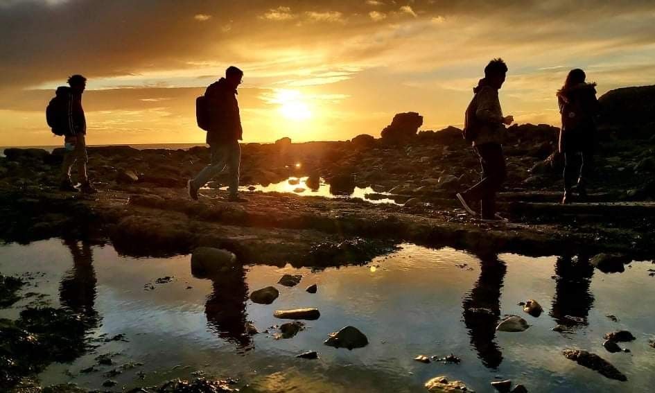 Four people walking through rocks and water in front of a sunset
