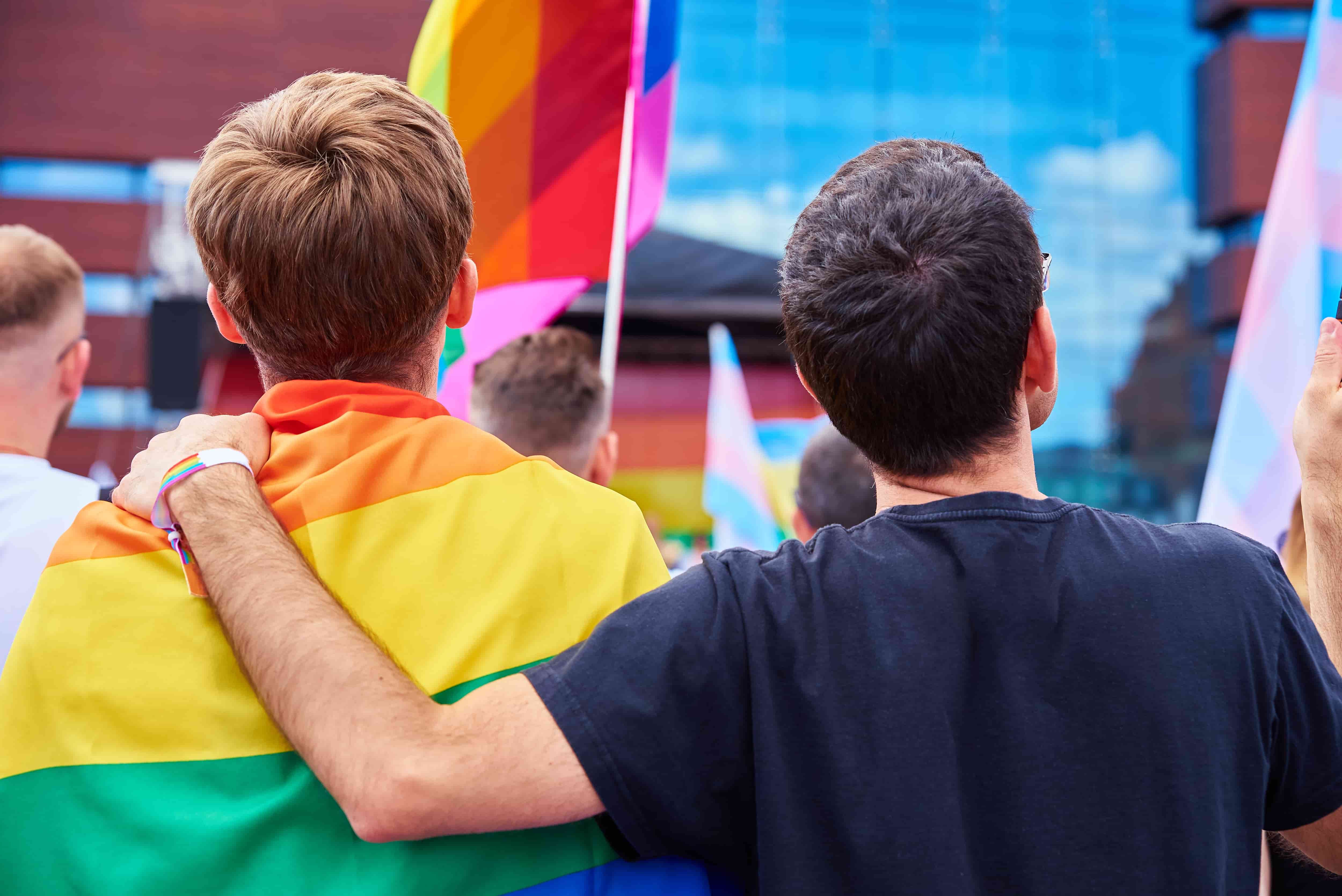 A man wearing a rainbow wristband puts his arm around the back of a man wrapped in a rainbow flag.