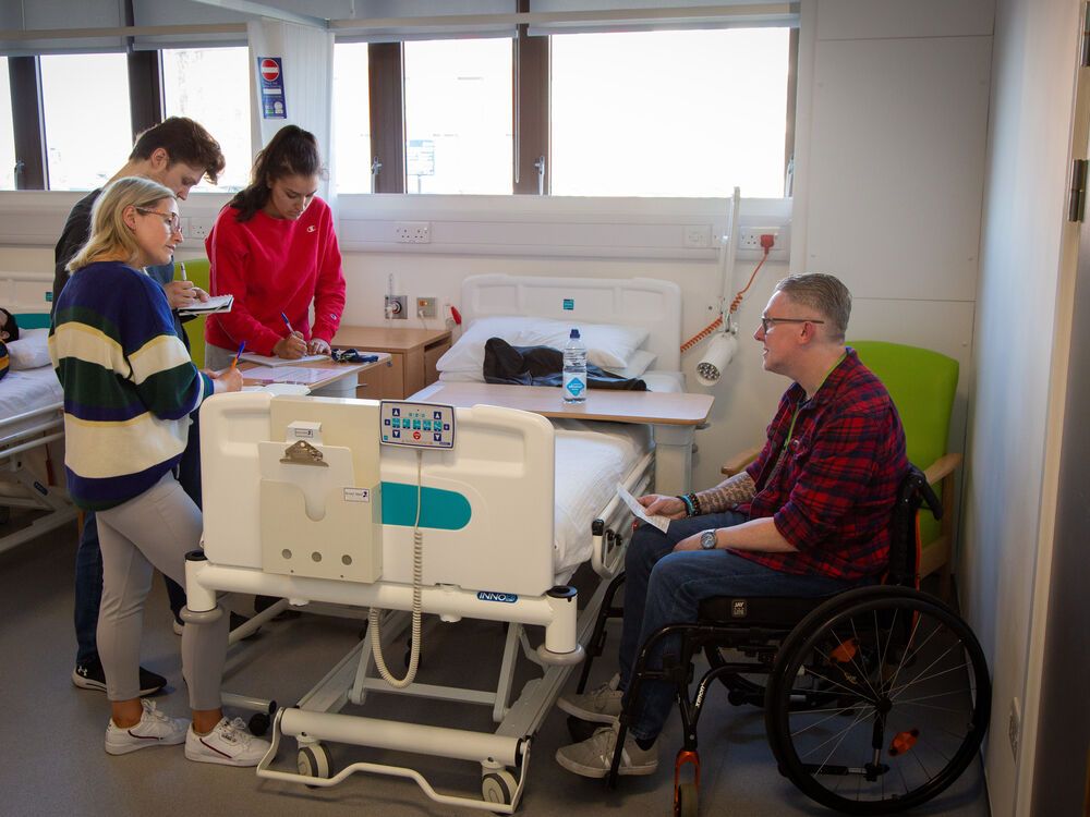 Three students working with a patient who is in a wheelchair during a clinical skills session