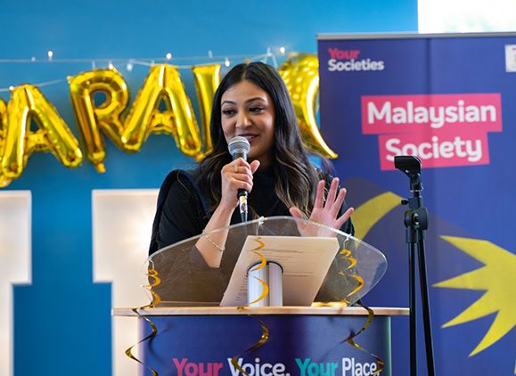 A student speaking into a microphone on stage at the Malaysian and Islamic Societies Eid celebration