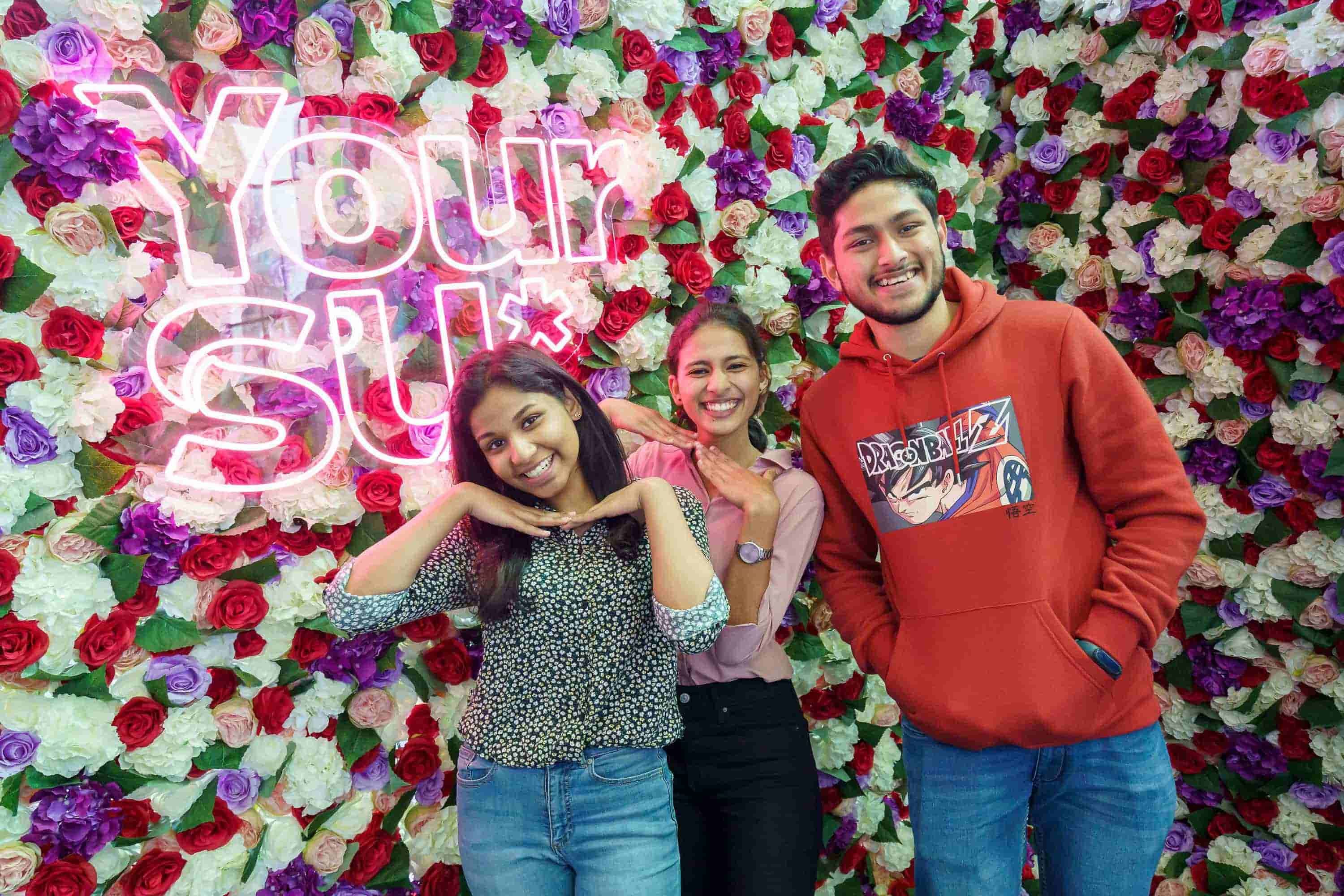 Three students standing in front of a flower wall with a sign that reads 