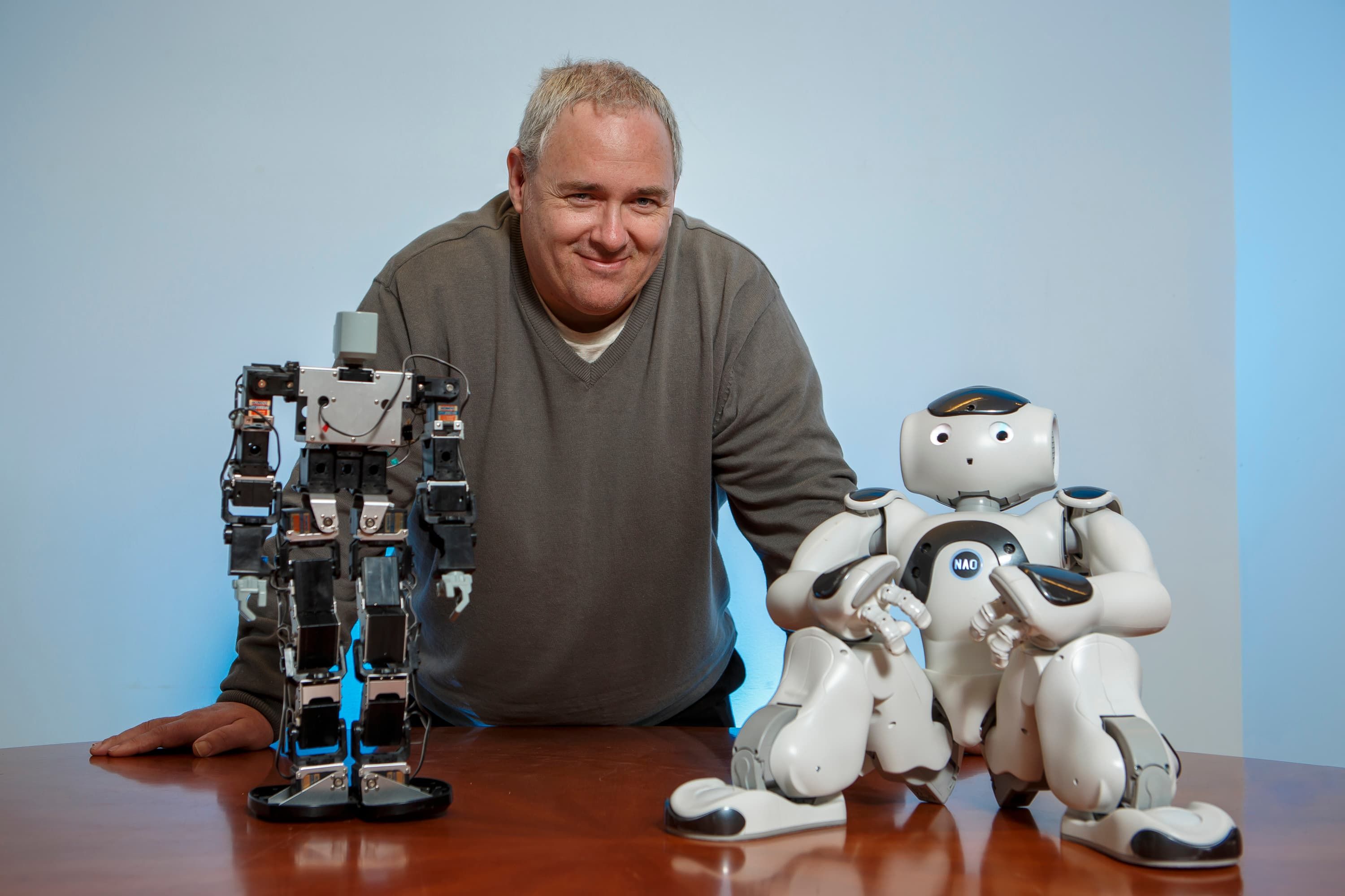 Professor John Murray smiling to camera next to a couple of small robots