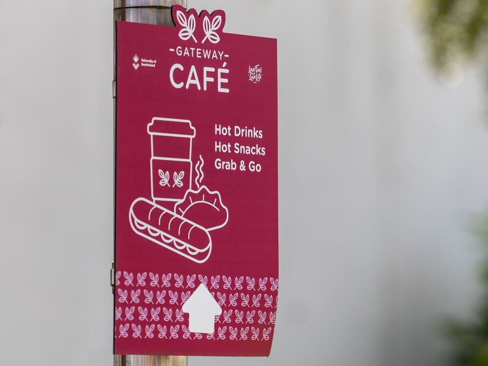 A pink sign on a post saying gateway cafe and hot drinks, hot snacks and food to go with a drawing of a sandwich and hot drink