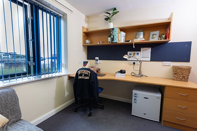 A desk with shelves above it and a desk chair with a University of Sunderland hoody draped over it. A small set of drawers and mini fridge are next to the desk.