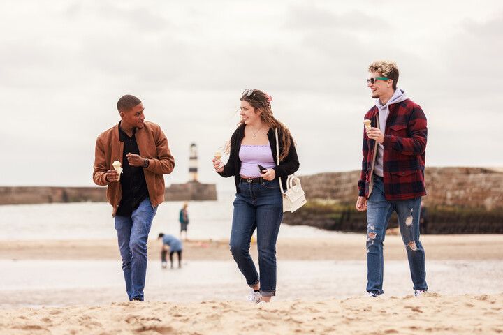 Three students walking together across a beach eating ice creams and talking