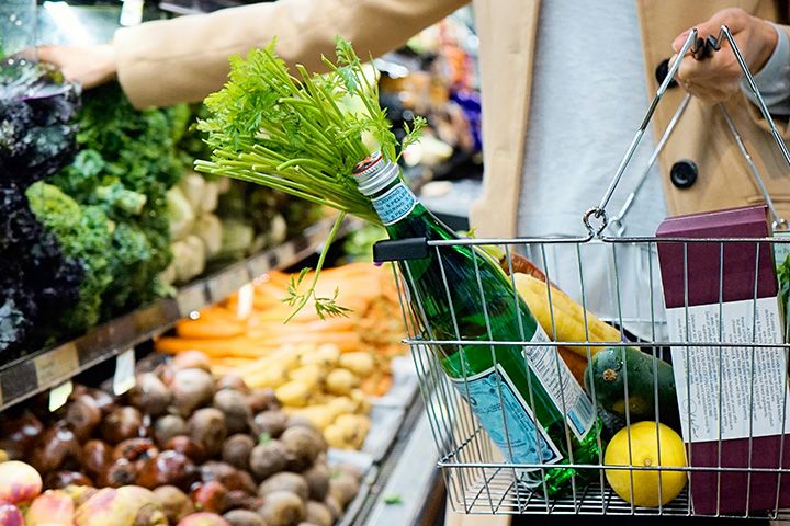 Hands holding a shopping basket full of groceries while reaching out to a shelf