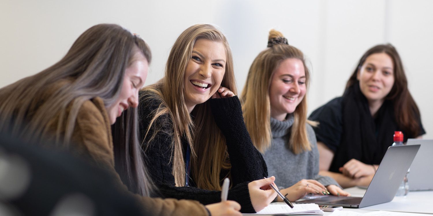 A group of female students are sitting together in a classroom, laughing at something off camera