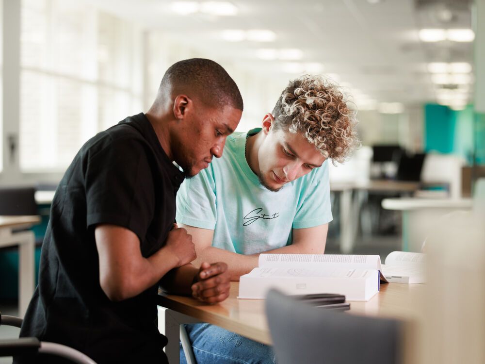 Two students sitting at a table in the library, looking at a textbook together