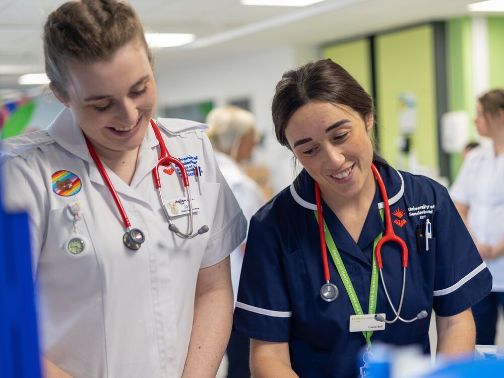 Two nursing students in uniform looking down at something that is off camera and smiling