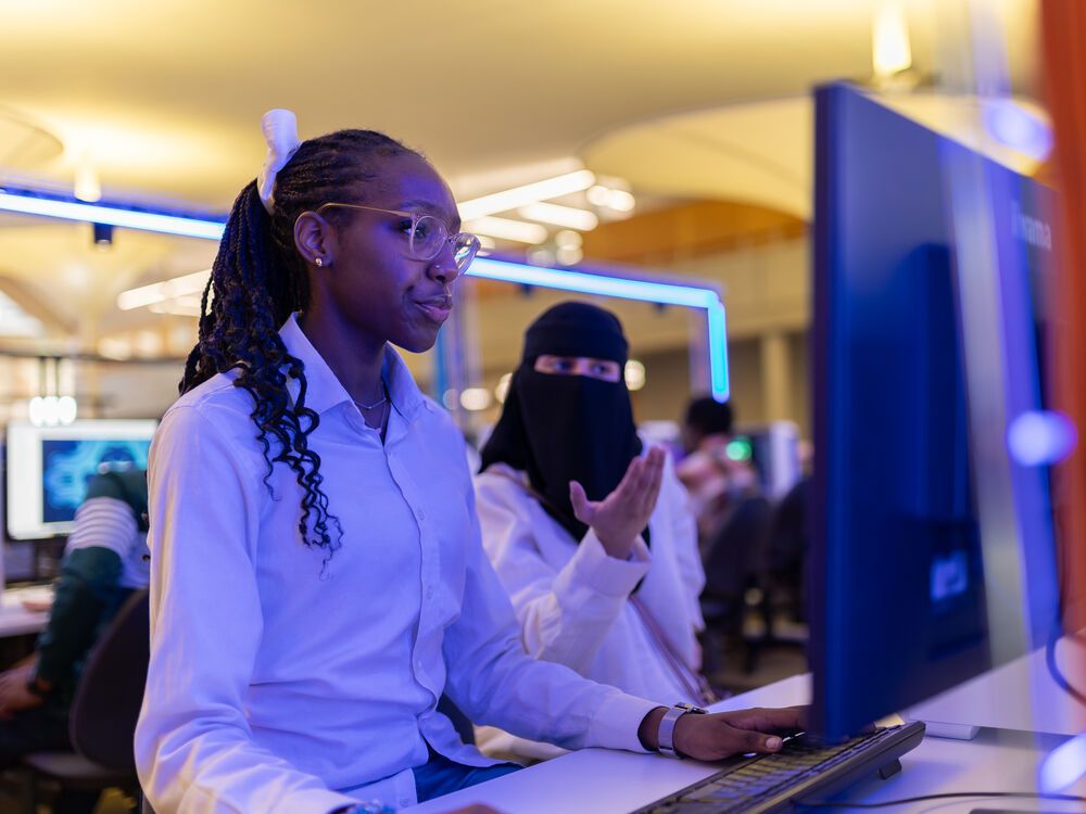 Two computer science students working together at a computer. One student is pointing at something at the screen while the other watches.