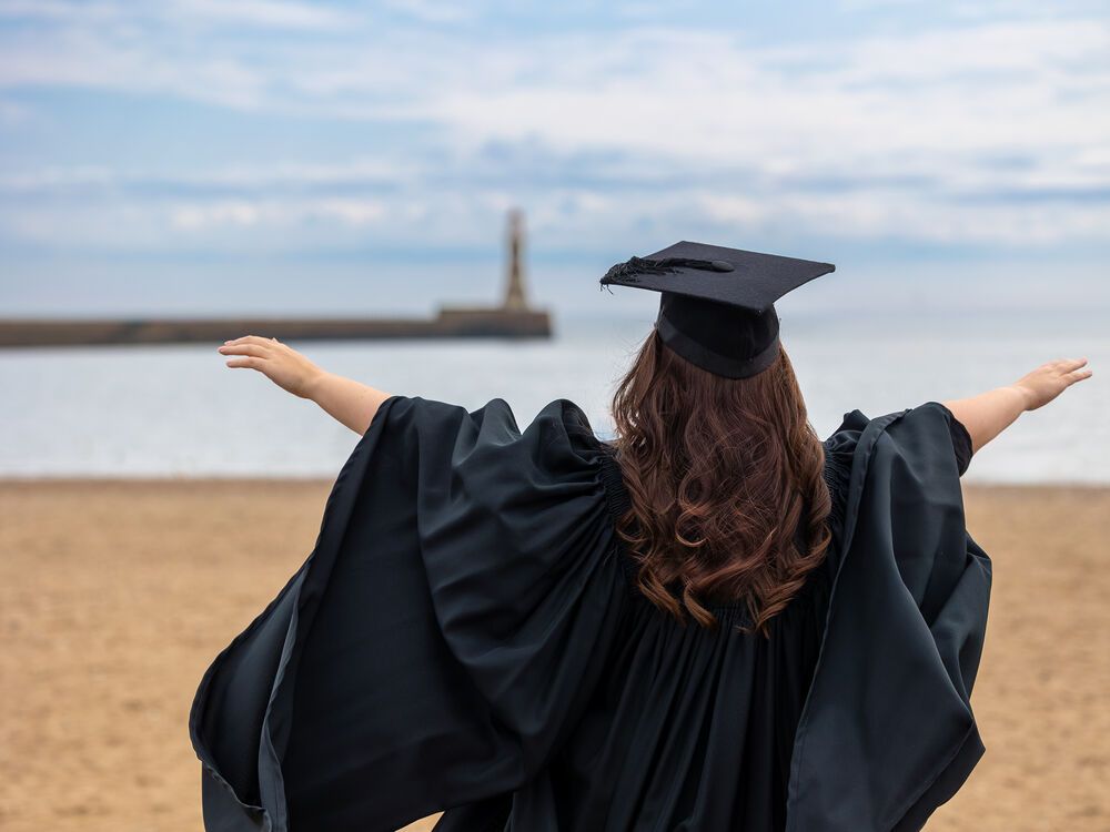 A graduate dressed in graduation ceremonial robes and mortar board spreading their arms in celebration on Roker Beach