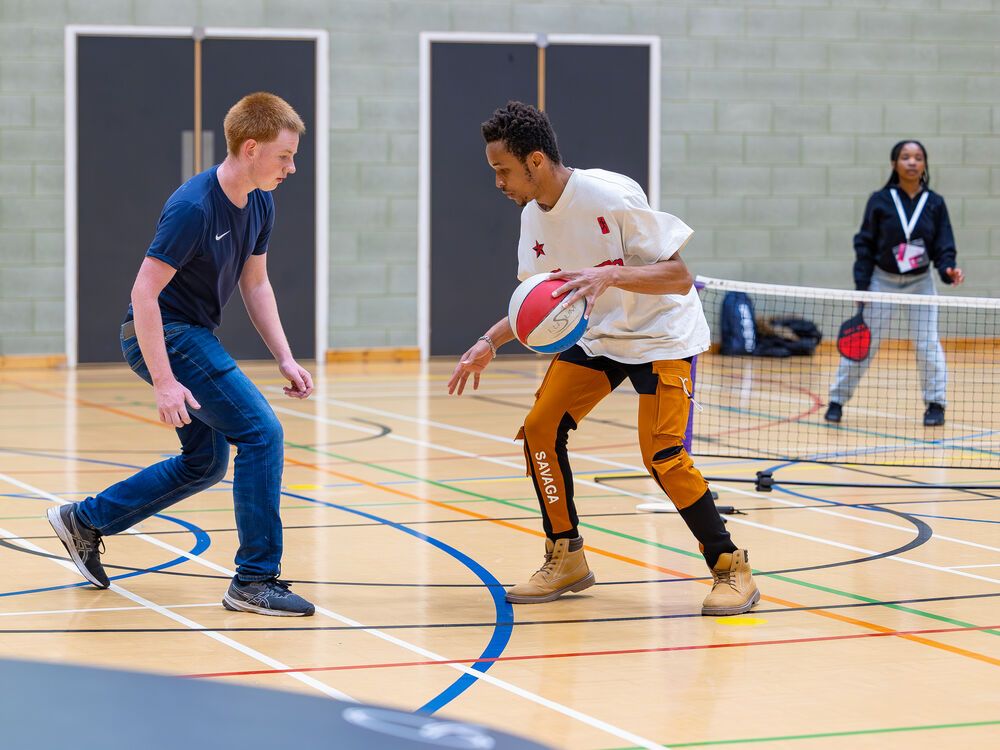 Two students playing basketball in the sports hall, while other students play other sports in the background