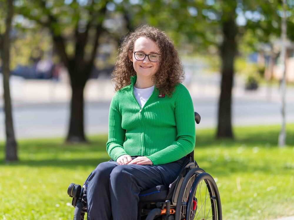Laura in a wheelchair smiling into the camera with trees and grass in the background