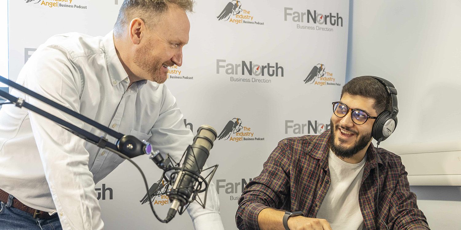 A student sitting in front of a microphone wearing headphones, smiling at another person