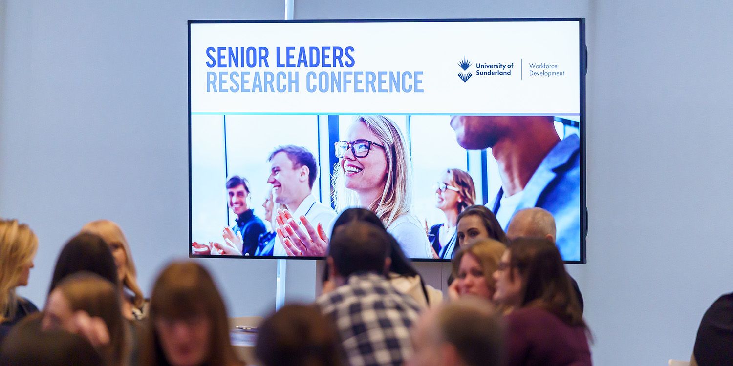 A large screen at the front of a room of people saying Senior Leaders Research Conference