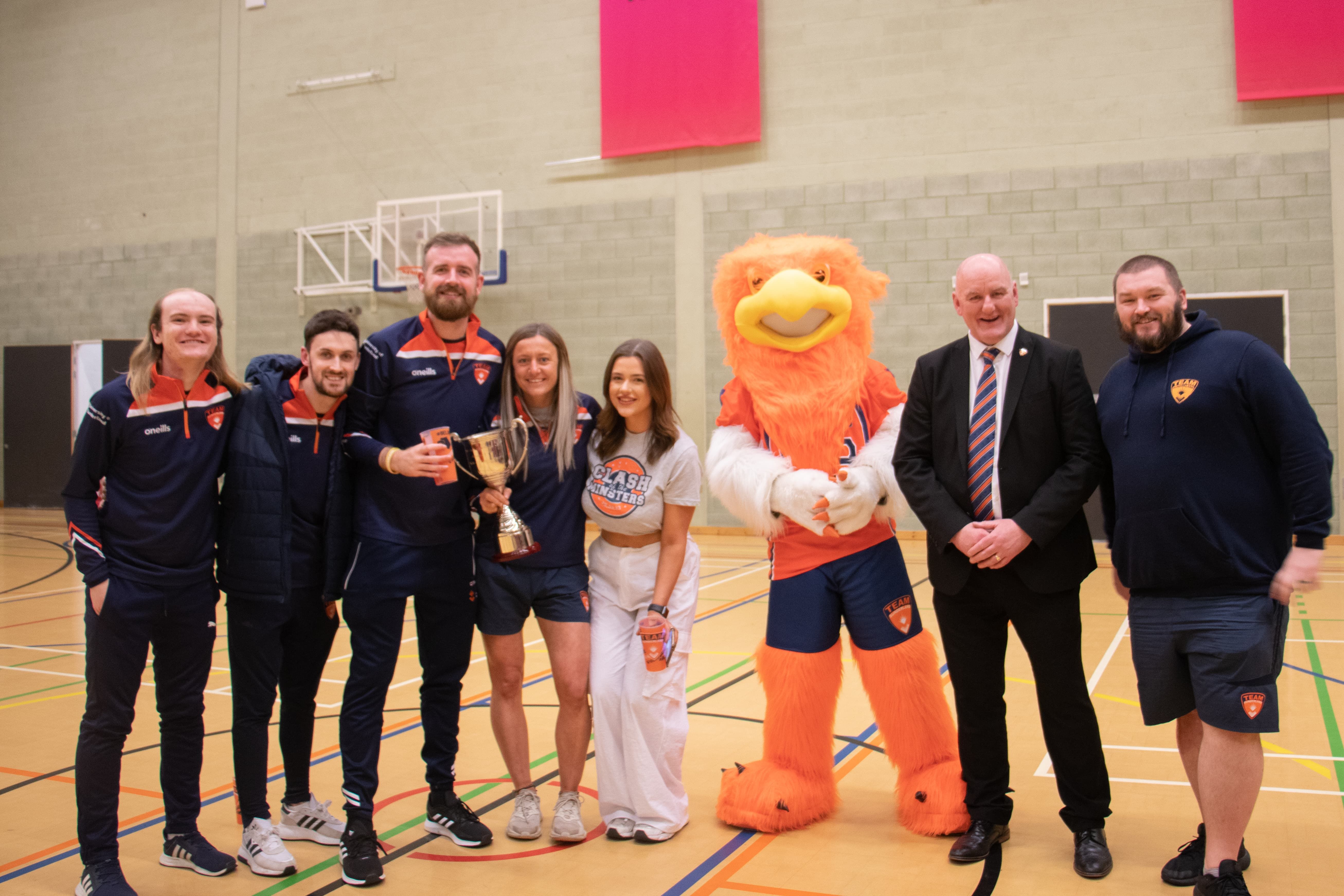 Team Sunderland standing in a line in the sports hall with the mascot (a griffin) and Sir David Bell, smiling