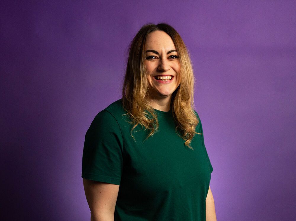 Helen stood facing the camera smiling with a purple background