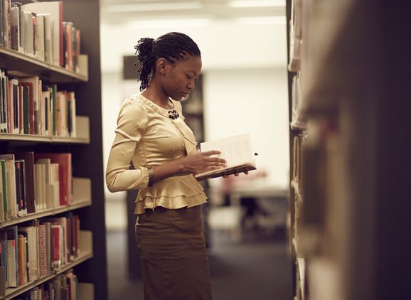 A student standing between the library shelves, looking at a book