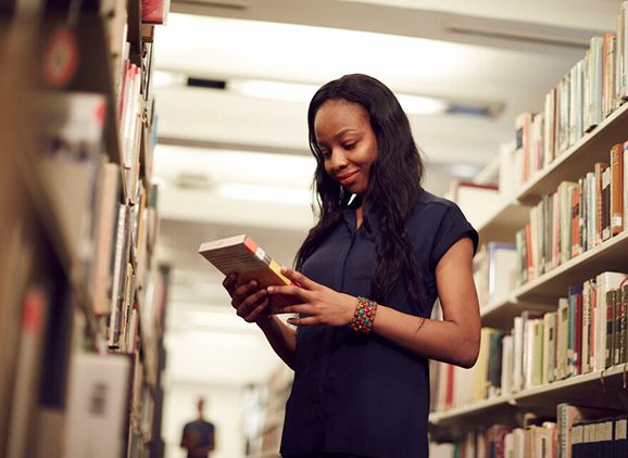 A student standing between the library shelves, looking at a book