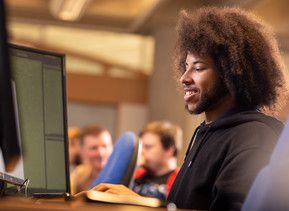 A student working at a computer, smiling. Two other students are visible in the background.
