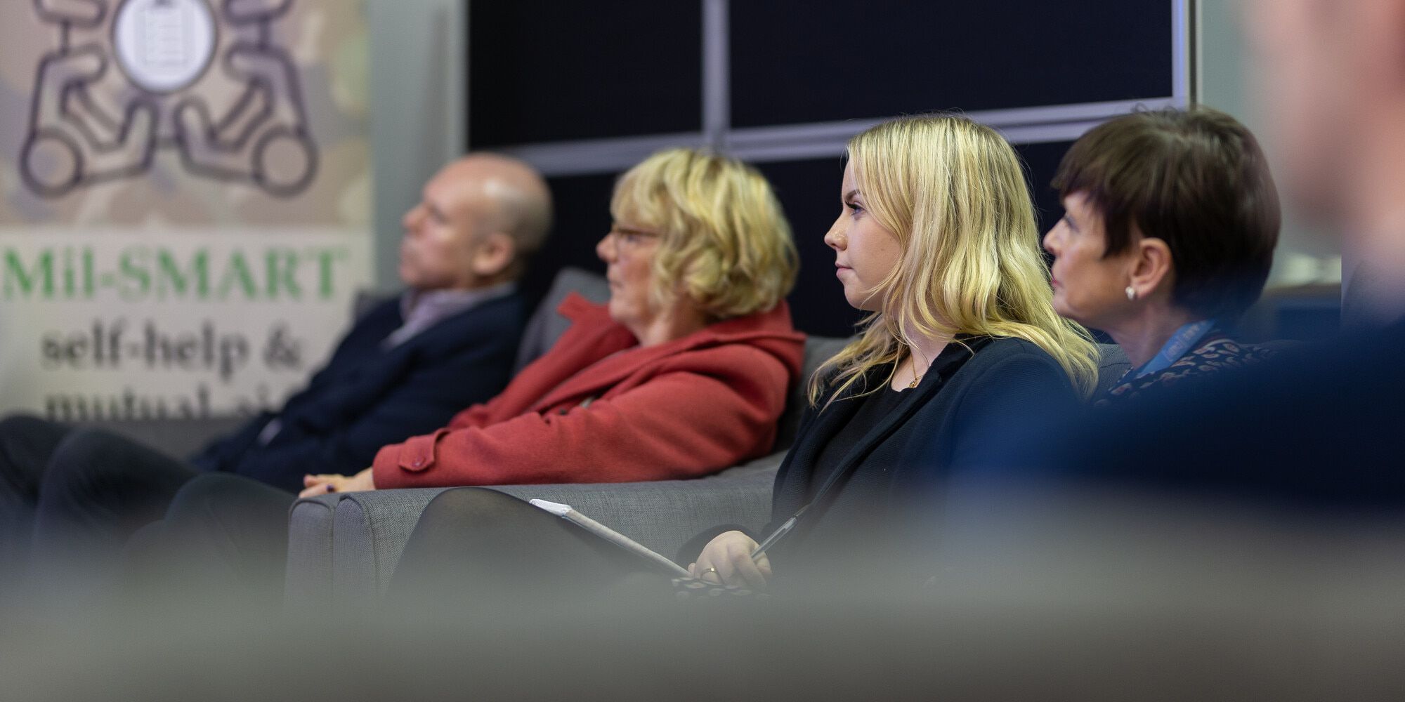 Four people sitting down on a sofa listening to and watching something off camera