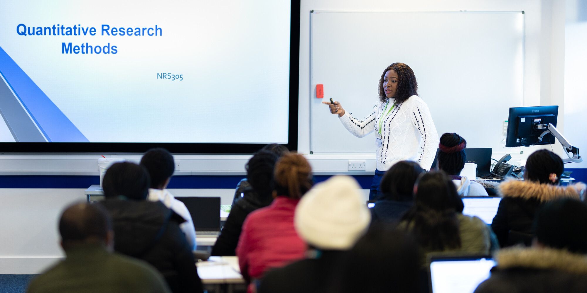 An academic standing at the front of a classroom, presenting to a class full of students who are listening