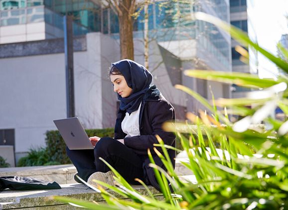 A student sitting cross-legged on the ground with a laptop, outside next to a leafy plant