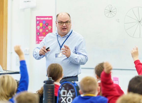 A male teacher standing at the front of a primary school classroom with children's hands in the air