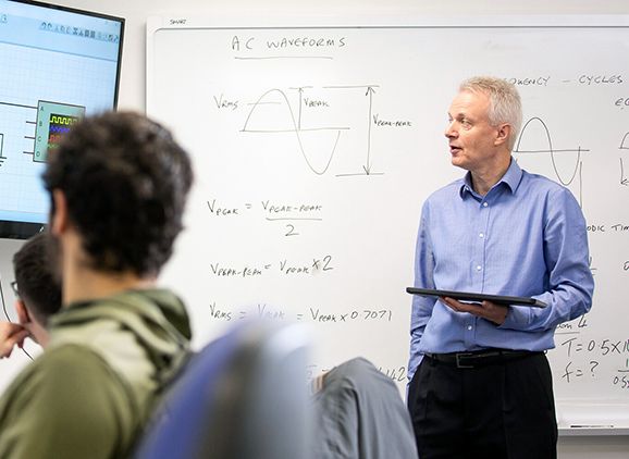 An academic teaching to a class with a whiteboard with equations behind him