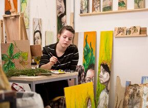 A student working in their studio surrounded by their paintings and other artwork