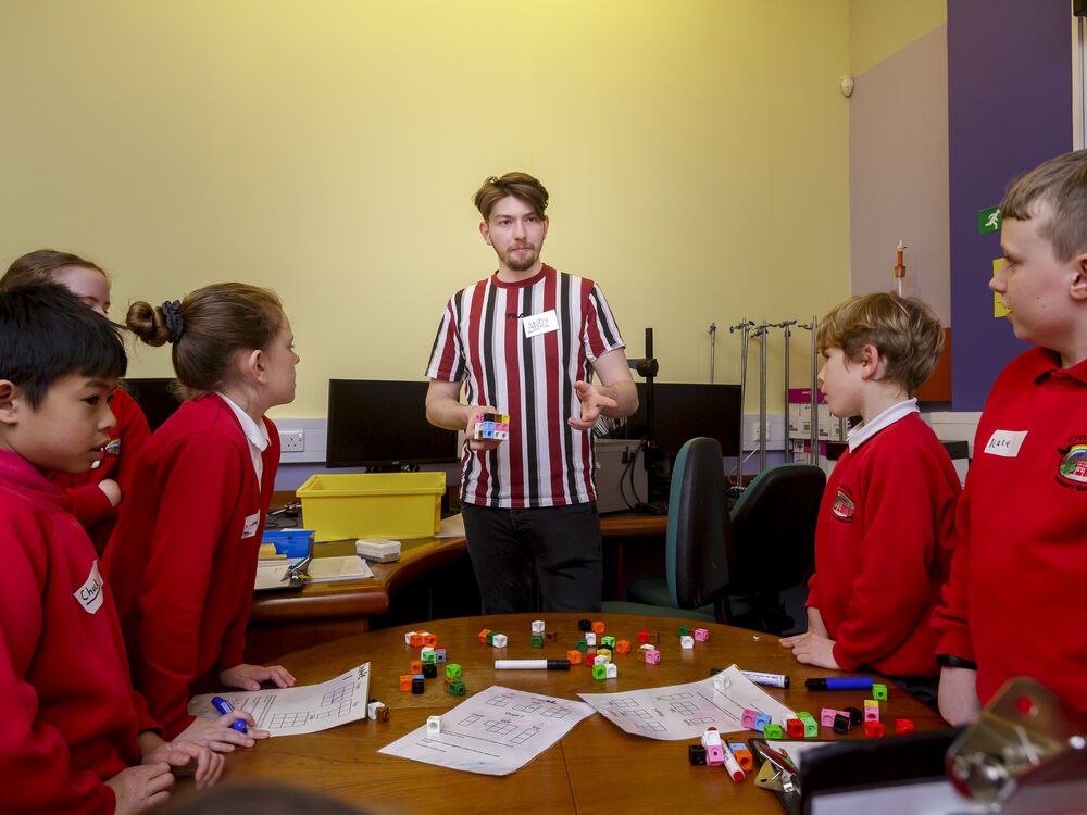 A student teacher standing at a table of school pupils demonstrating a maths problem with small cubes