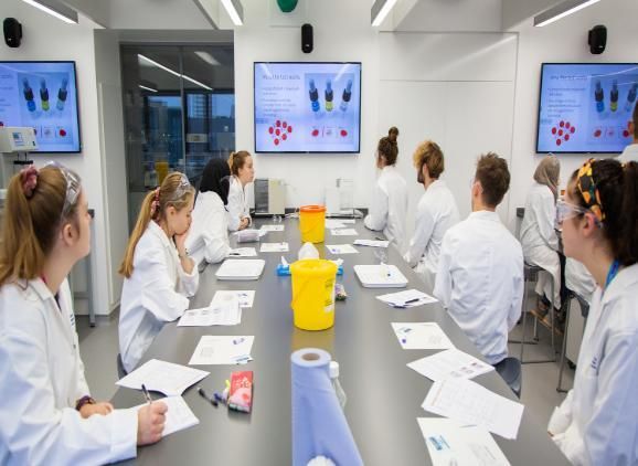 A group of Medicine students are sitting in white lab coats at a bench in a laboratory, facing away from the camera and looking at a TV screen