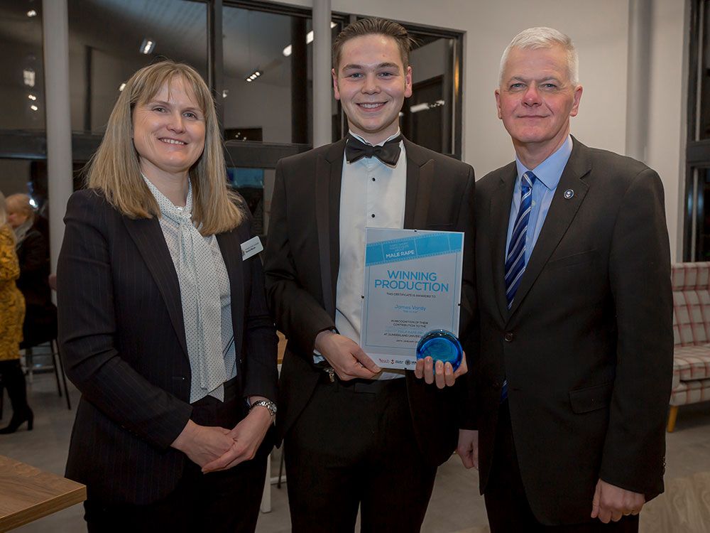 A student holding a certificate and trophy, standing alongside the Vice Chancellor and a client, smiling towards the camera.