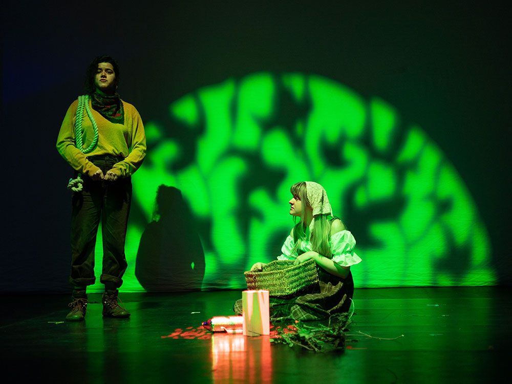 Two students performing in a production. One sits on the floor holding a basket, looking up to the other student who is standing and looking into the distance.