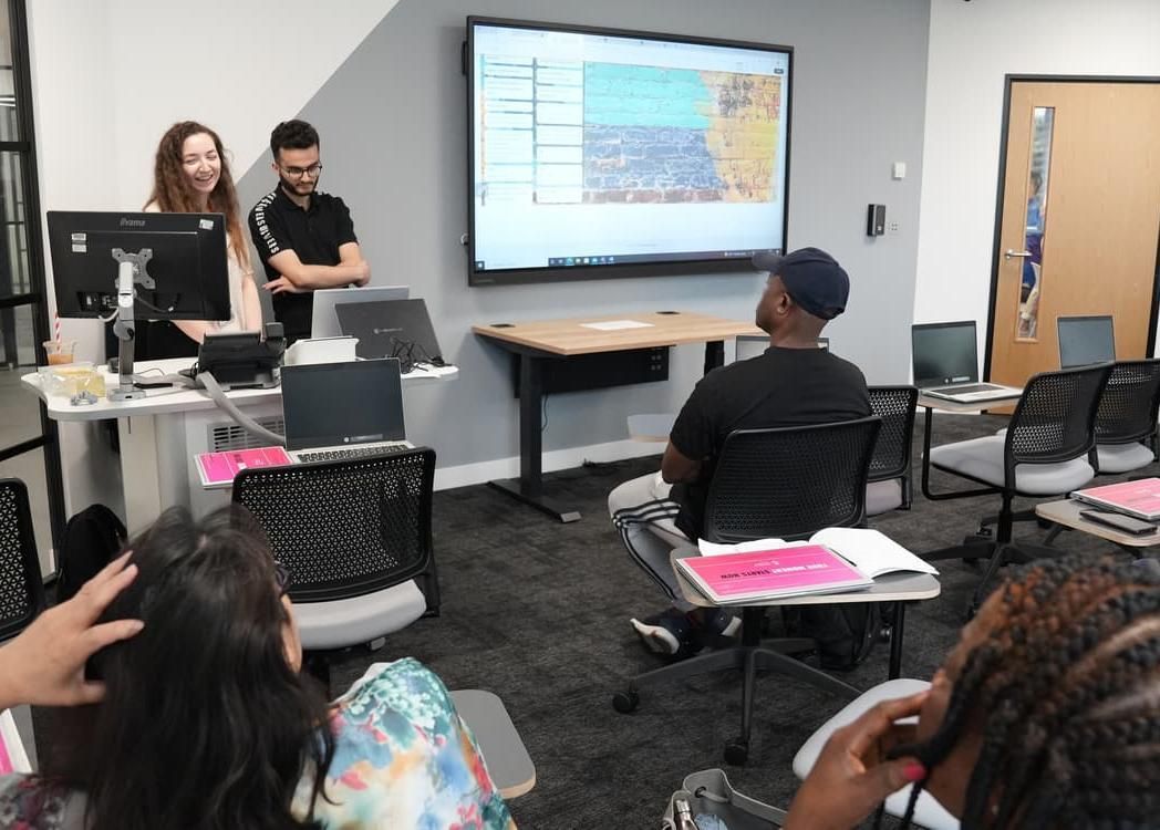 Two students giving a presentation at the front of their class using laptops and a projector to their peers