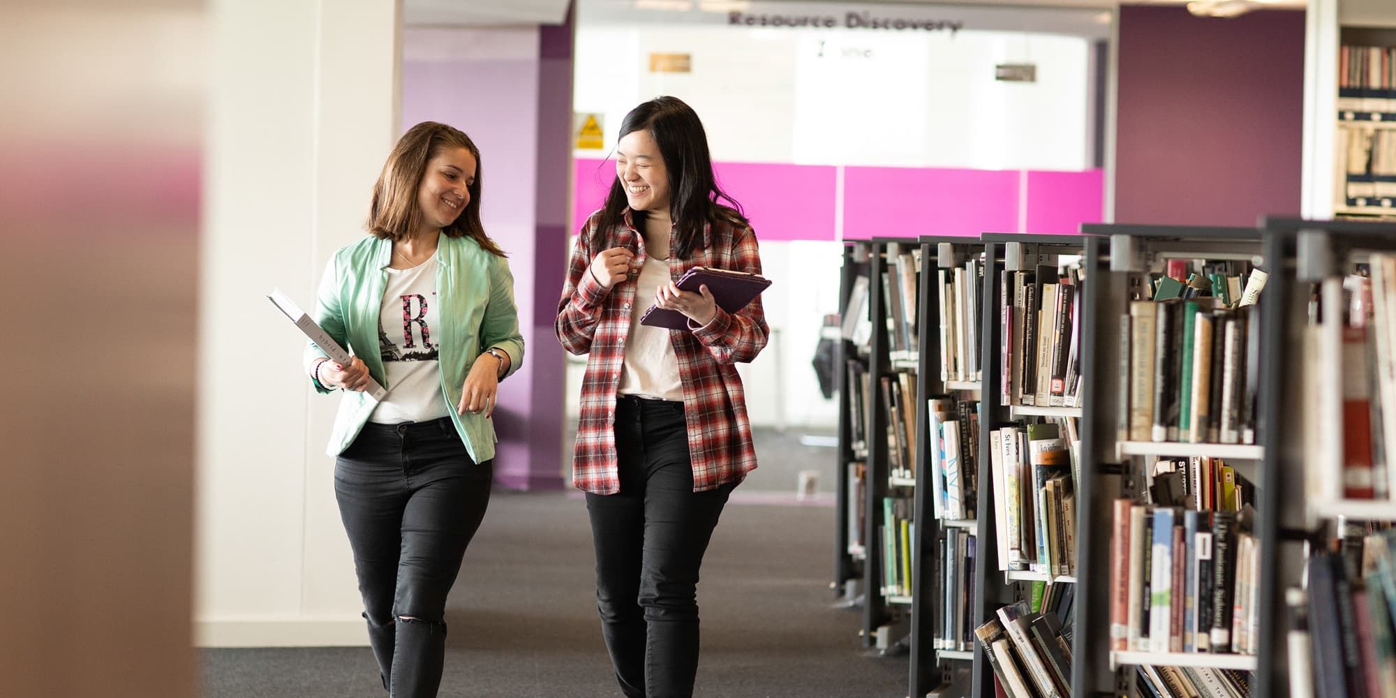 Two students walking through the library with books laughing together