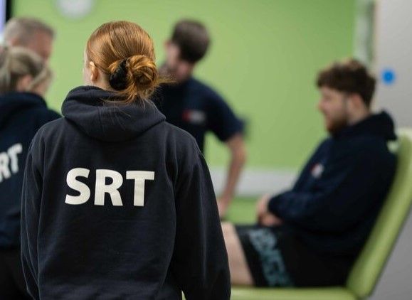 Students with SRT written on the back on their tops gathering around a student on physio bed