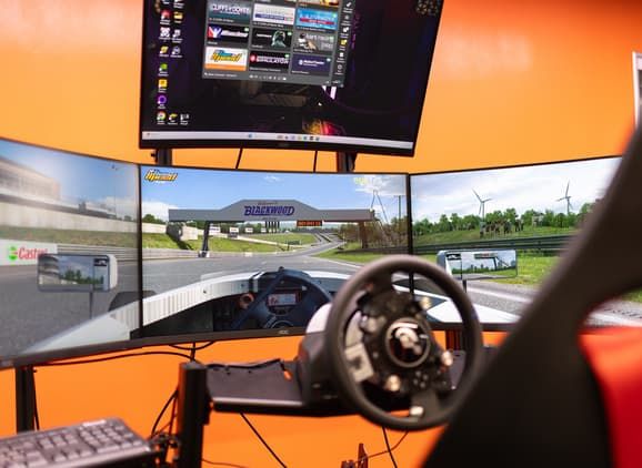 Three screens with a road track on with a steering wheel and gaming chair infront of it