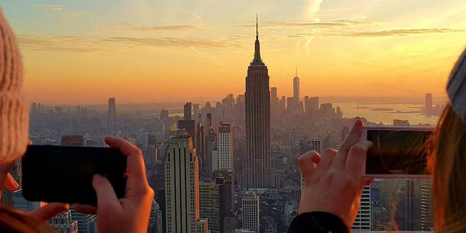 A view of the New York city skyline with two students taking a photo of the Empire State Building on their phones